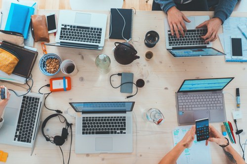 Tavolo visto dall'alto, adibito a un lavoro di gruppo, su cui si vedono pc, telefoni, penne, materiale di cancelleria e le braccia/mani delle persone coinvolte